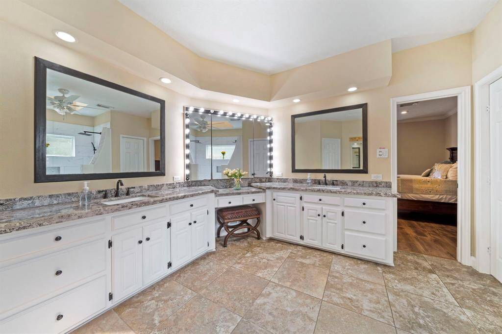 This spacious primary bathroom features dual sinks with granite countertops, lots of storage with white cabinetry, and large mirrors enhanced by soft lighting. The room is brightly lit, with tiled floors.