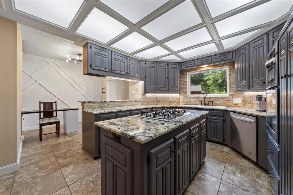 This kitchen features a central island with a gas cooktop, granite countertops, and cabinetry. It offers ample storage, a large window for natural light, and modern appliances, creating a functional and stylish cooking space.