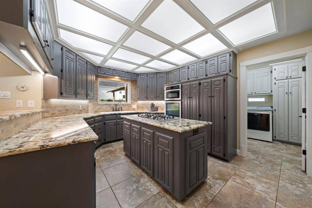 This spacious kitchen features elegant gray cabinetry, granite countertops, and a center island with a built-in cooktop. The layout offers generous storage and an adjacent laundry room, while large ceiling lights provide ample illumination.