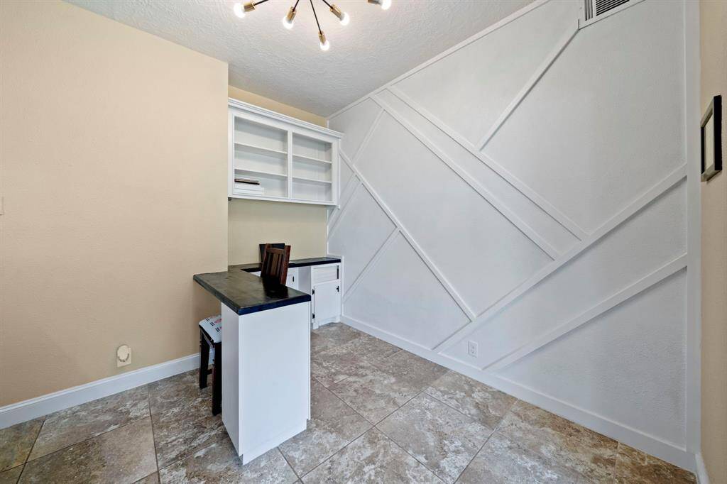 This room features a modern design with geometric wall detailing and tiled flooring. It includes built-in shelving, a desk area with a dark countertop, and a stylish light fixture, making it ideal for a home office or study space.