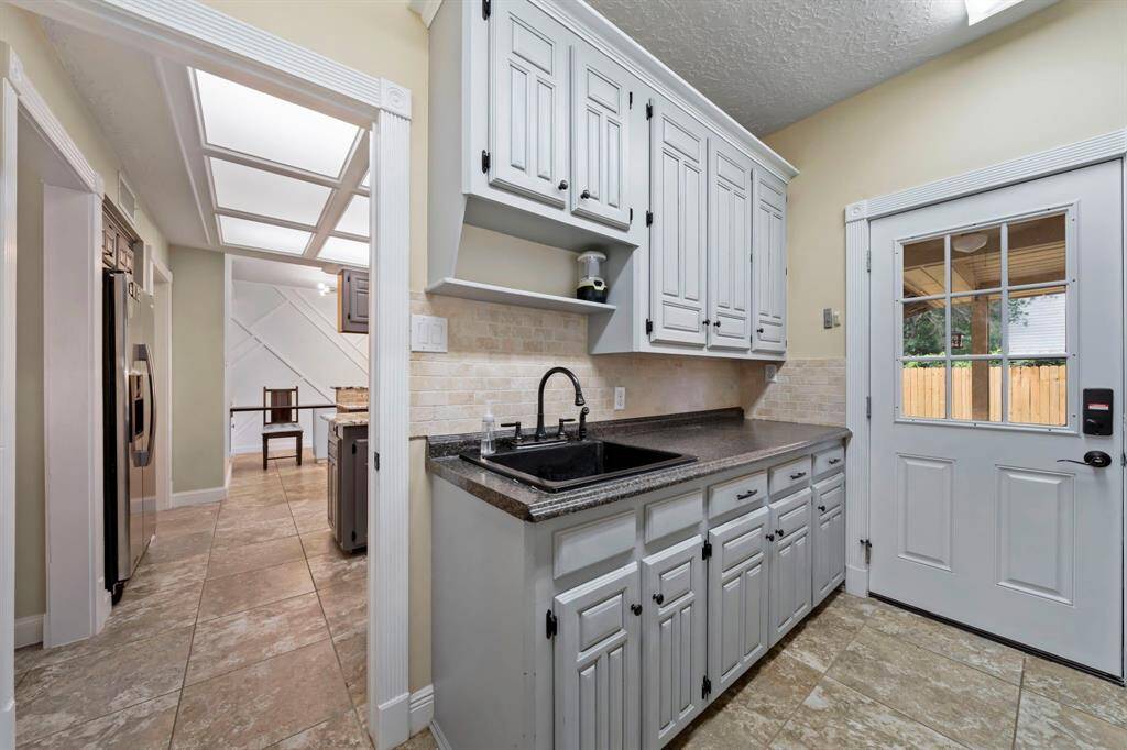 The laundry room has lots of cabinetry, a dark countertop, and a tiled backsplash. The room is well-lit with natural light coming through a windowed door that leads outside. The laundry room has a modern, clean design with ample storage space.