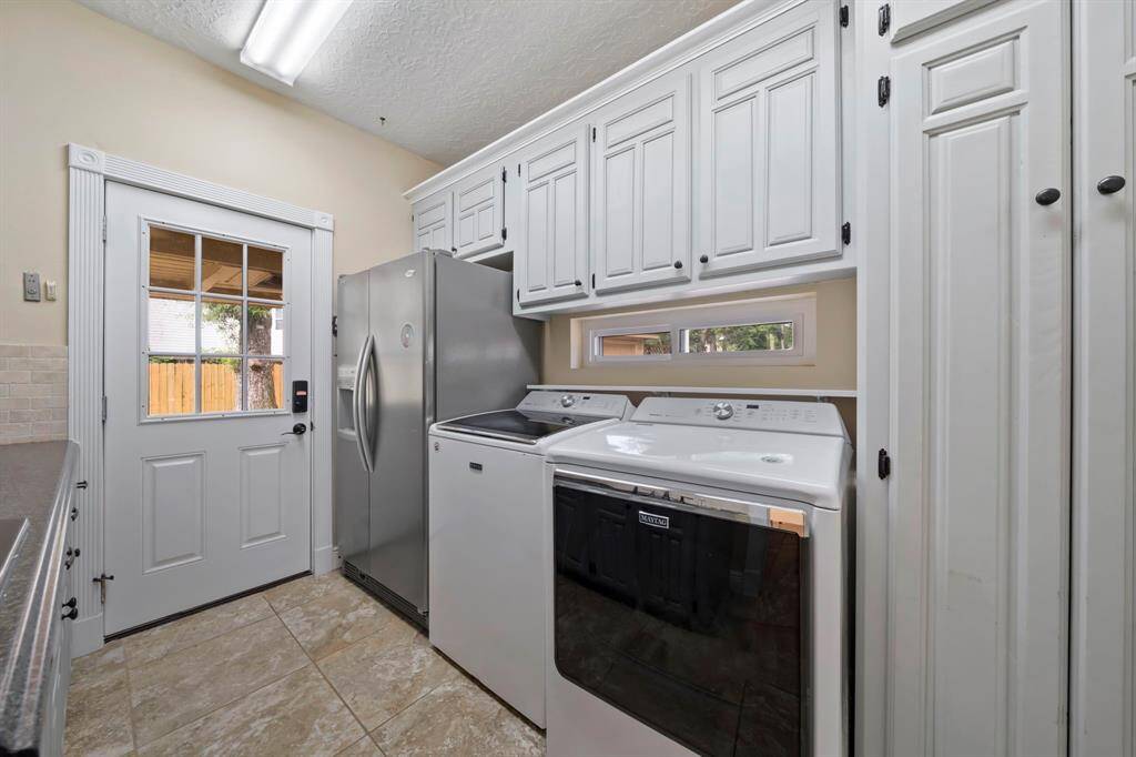Another view of the laundry room shows just how bright and functional this room is featuring lots of cabinetry, a washer, dryer, a stainless steel refrigerator, and tiled flooring. A door with a window provides natural light and outdoor access.