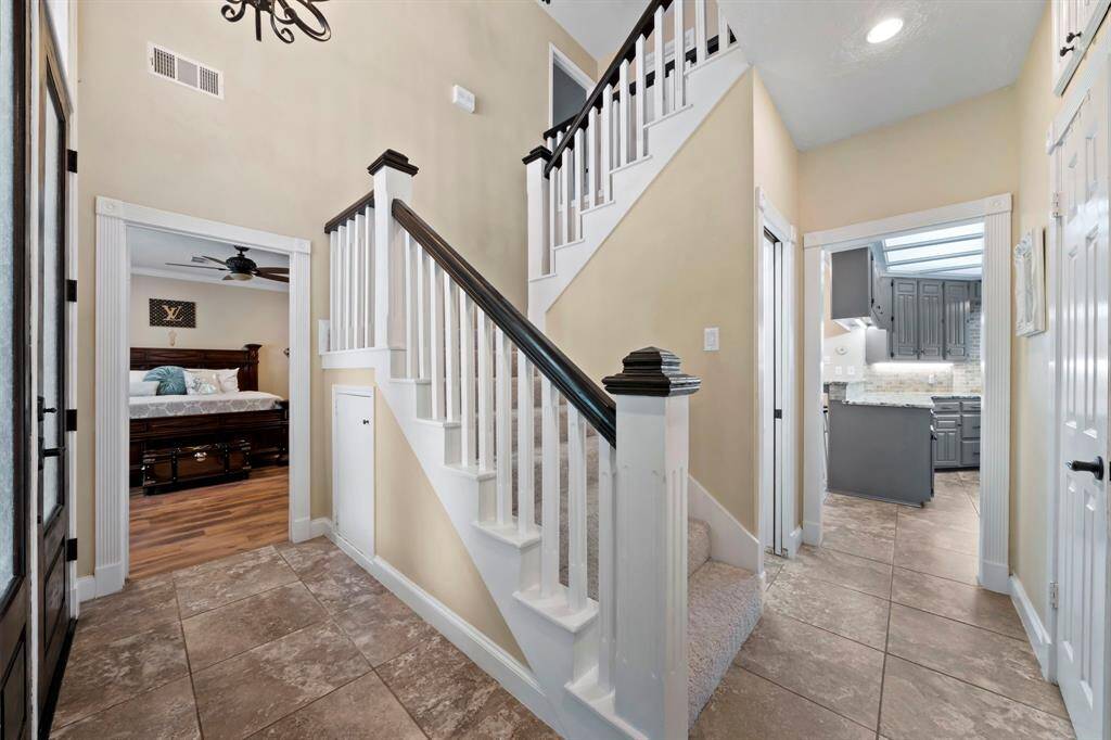 The entryway with tiled flooring, leads to the staircase. The space opens into a huge primary bedroom with wood-like flooring on the left and a modern kitchen with gray cabinetry on the right.