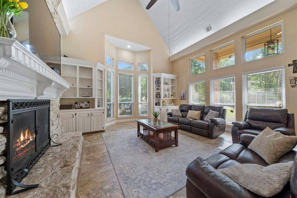 The living room features high ceilings, large windows offering abundant natural light, a cozy stone fireplace, and elegant solid wood built-in shelving and recess lighting. The open layout is perfect for relaxing and entertaining.
