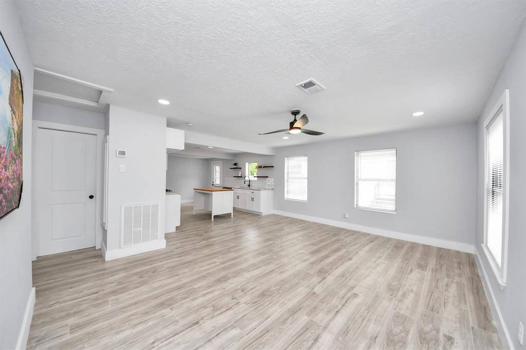 This photo shows a bright, open-concept living space with light wood flooring and neutral walls. It features a modern ceiling fan, recessed lighting, and large windows that let in plenty of natural light. The area seamlessly connects to a stylish kitchen with white cabinetry and a central island.