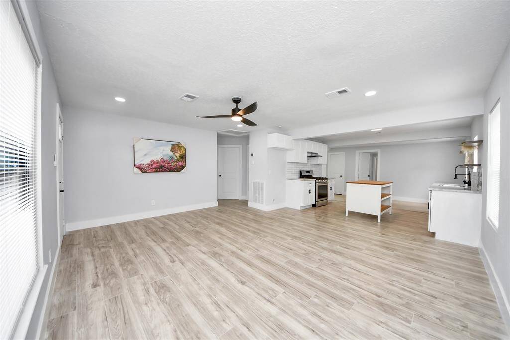 This bright, open-concept living area features modern light wood flooring and a ceiling fan. The adjacent kitchen offers sleek white cabinetry, a stainless steel stove, and ample counter space, perfect for entertaining and daily living.