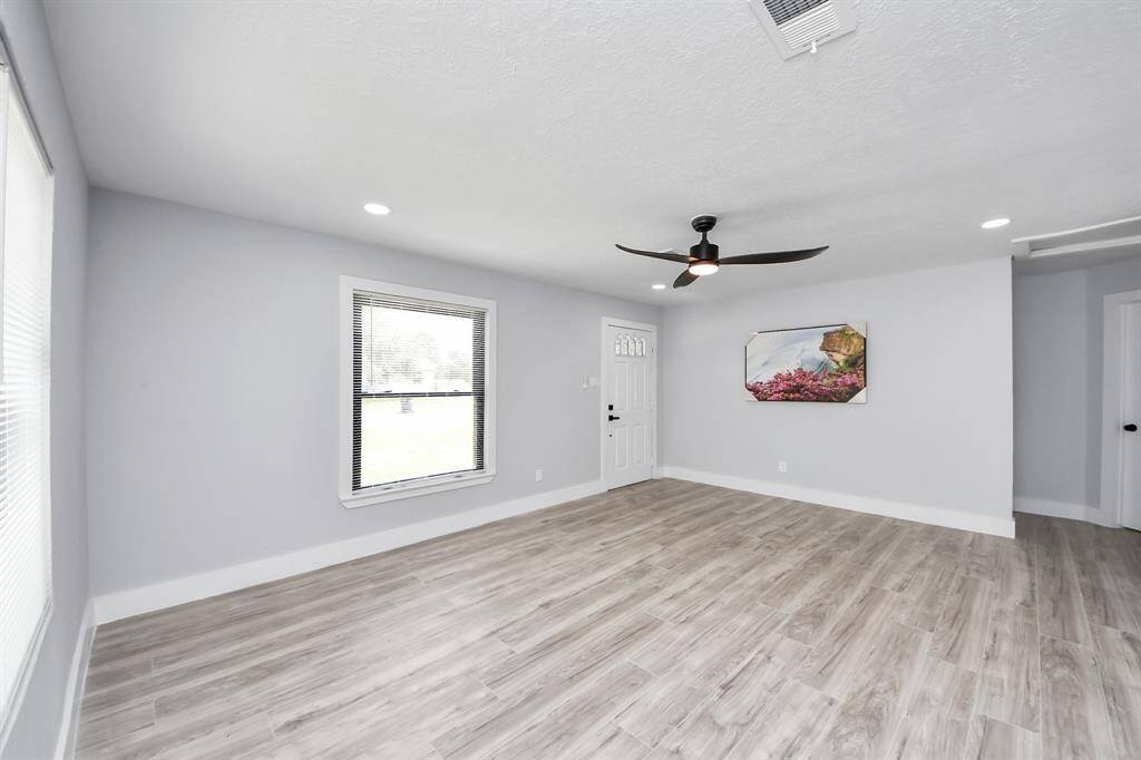 This spacious, bright living area features modern wood-style flooring, a ceiling fan, and ample natural light from large windows. The neutral walls and recessed lighting create a welcoming and versatile space.