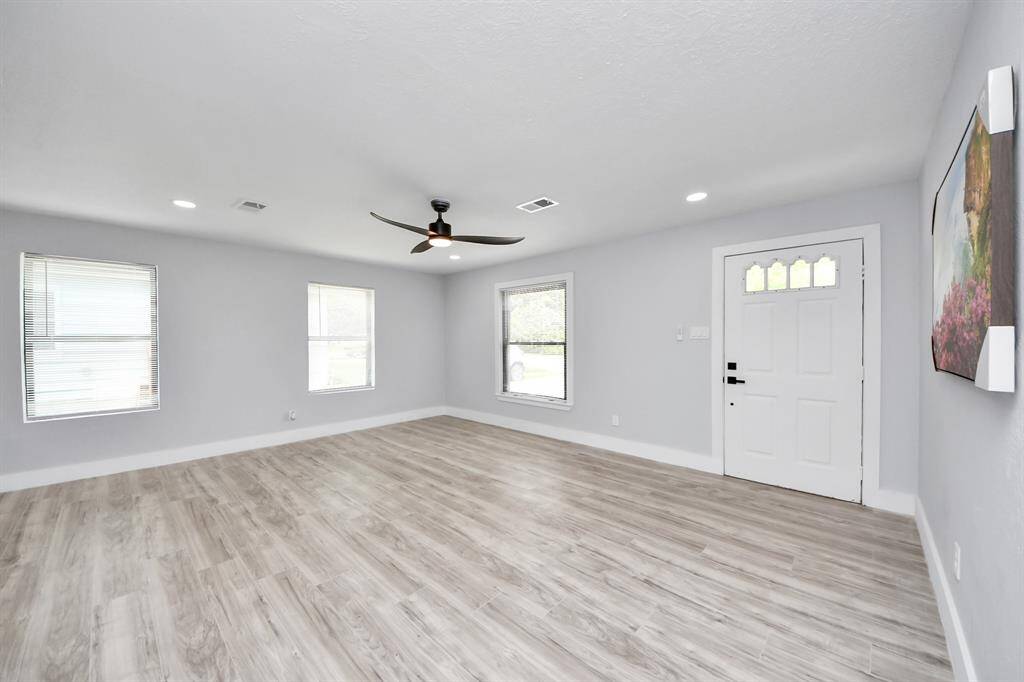This spacious and bright living room features light wood flooring, neutral walls, and ample natural light from three large windows. It includes recessed lighting, a modern ceiling fan, and a stylish front door, creating a welcoming and airy atmosphere.