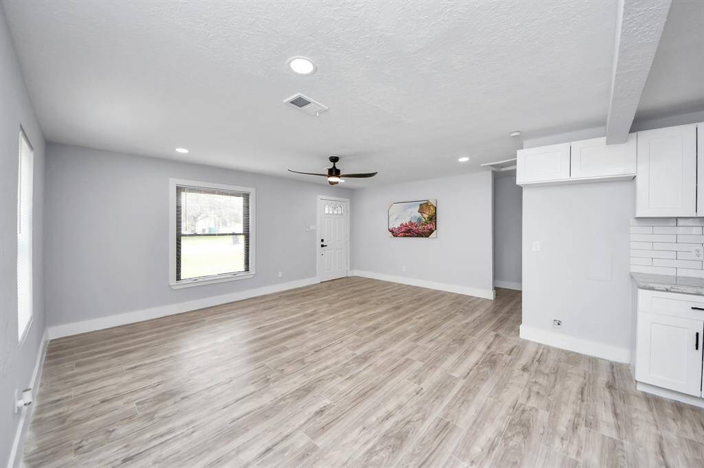 This spacious, open-concept living area features modern laminate flooring, neutral walls, and abundant natural light. It includes a ceiling fan, recessed lighting, and a kitchen with white cabinets and subway tile backsplash, creating a bright, inviting atmosphere.