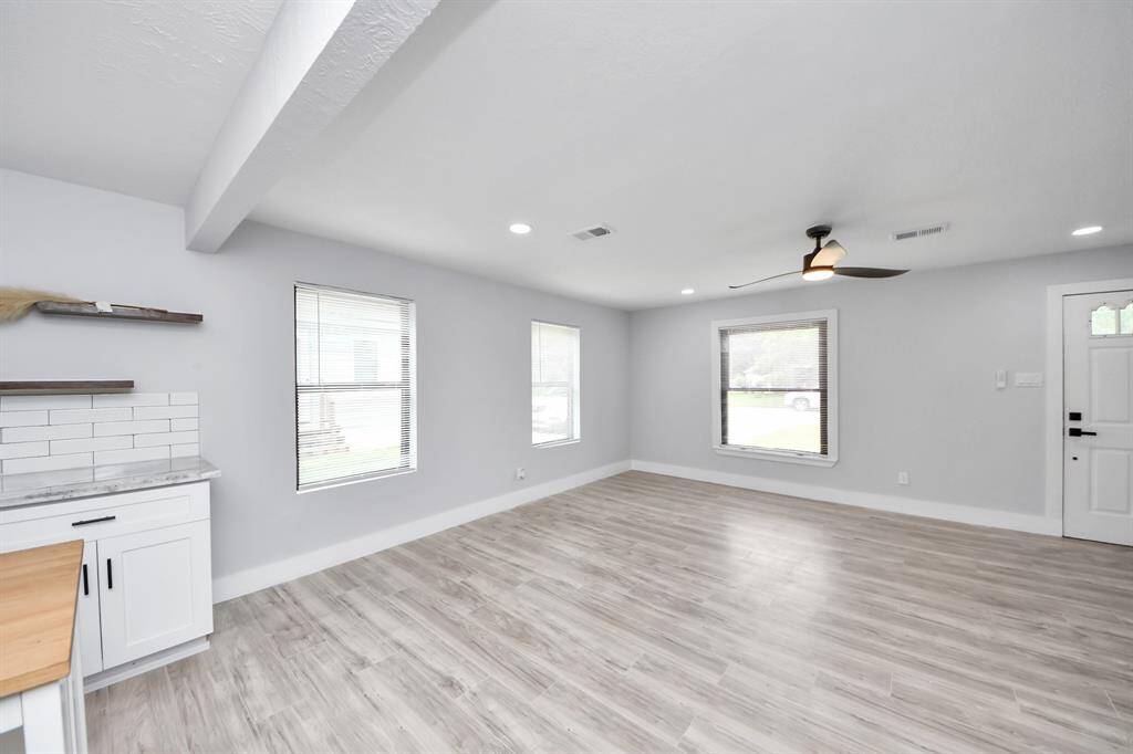 Bright and spacious open living area with light wood flooring, modern ceiling fan, and large windows. Freshly painted walls and a partial view of the kitchen with white cabinetry.