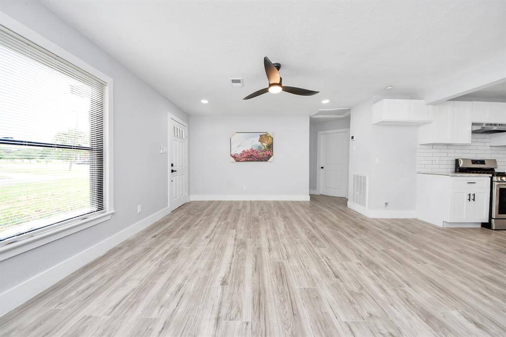 This photo showcases a bright, open-concept living space with light wood flooring and fresh white walls. It features a modern ceiling fan, large window for natural light, and an adjacent kitchen with stainless steel appliances and white cabinetry.