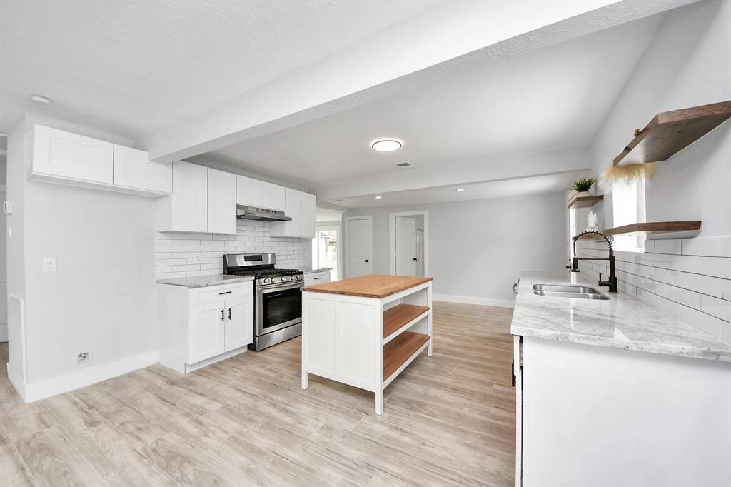 This kitchen features modern white cabinetry, a stainless steel stove, and a subway tile backsplash. It includes a central island with wooden shelves and a spacious sink area with marble-like countertops. The space is bright with ample natural light and wood-look flooring.
