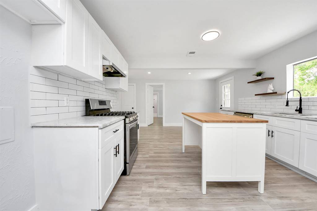 This modern kitchen features sleek white cabinetry, a central island with a wooden countertop, and stainless steel appliances. The space is bright with a large window, open shelving, and a stylish subway tile backsplash.
