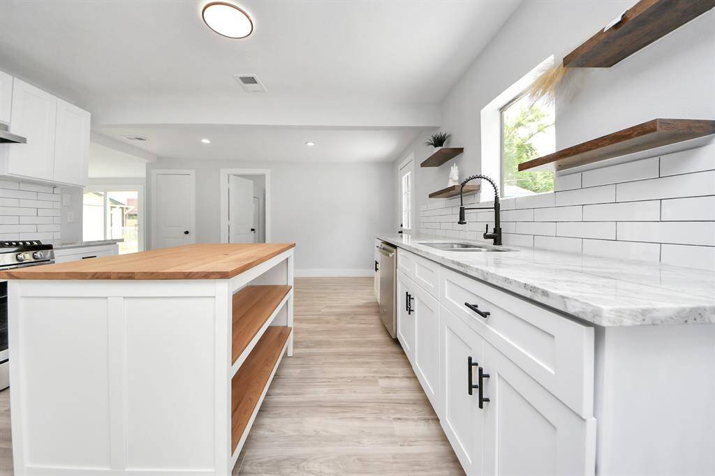 This kitchen features a modern design with white cabinetry, a central island with wood countertops, and sleek stainless steel appliances. The space is brightened by natural light and includes open shelving and a stylish subway tile backsplash.