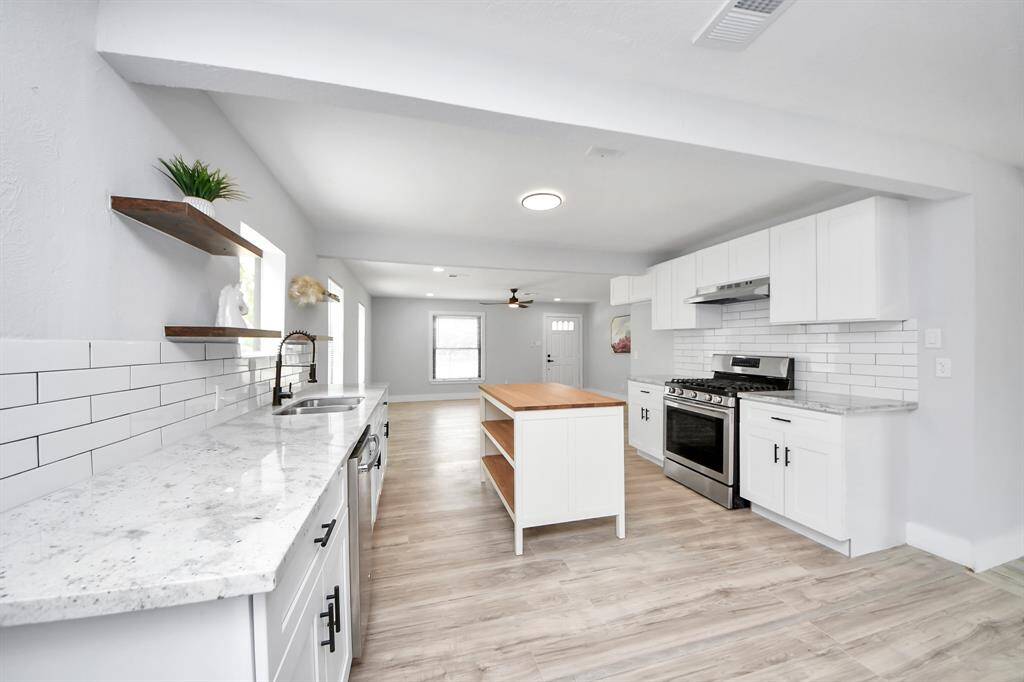 Bright, modern kitchen with open layout. Features white cabinetry, subway tile backsplash, stainless steel appliances, and a central island with wood countertop. Light wood flooring and ample natural light enhance the airy feel.