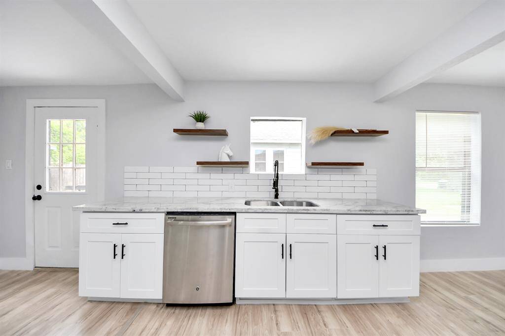 Bright, modern kitchen with white cabinetry and marble countertops. Features a double sink, stainless steel dishwasher, and open shelving. Natural light through a window and adjacent door enhance the airy feel.
