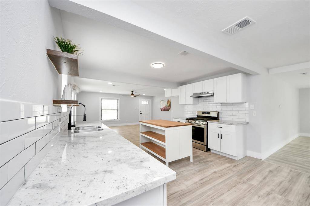 This bright, modern kitchen features white cabinetry, stainless steel appliances, and a central island with a wooden countertop. The space is open and airy, with light wood flooring and ample natural light.