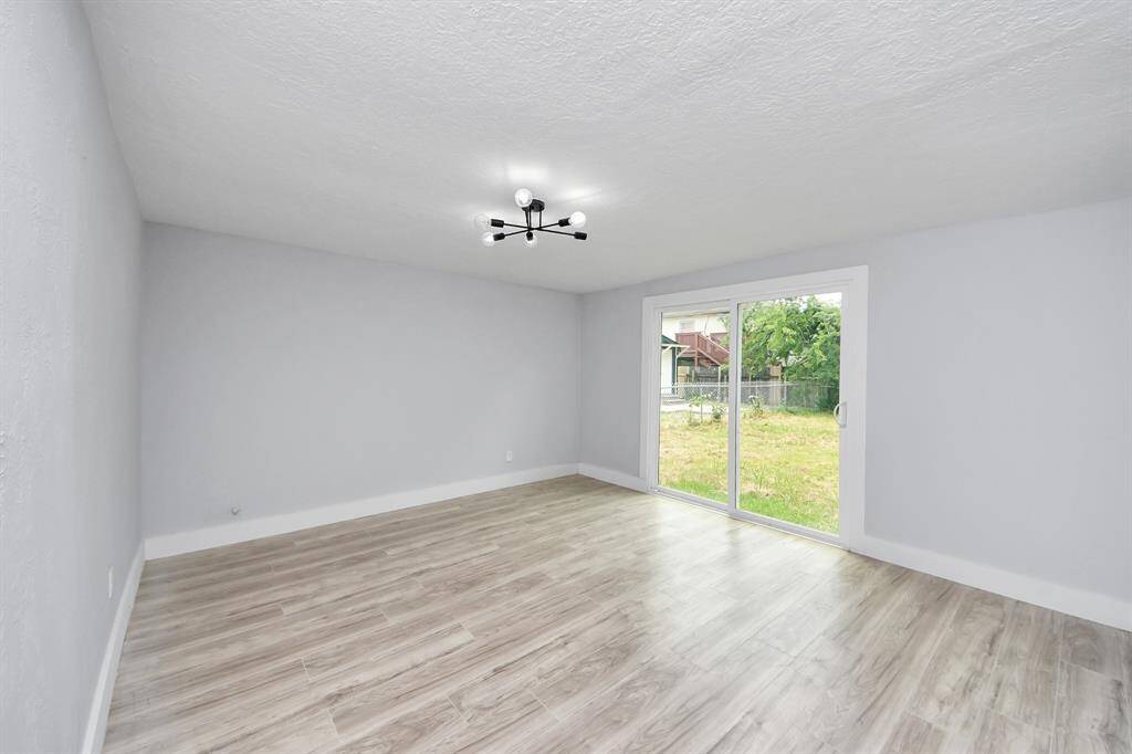 This room features light wood flooring, a modern ceiling light, and large sliding glass doors that open to a backyard, providing ample natural light and a view of greenery.