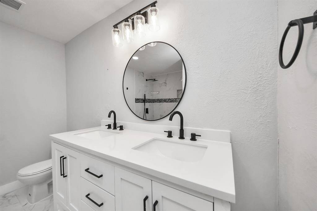 This bathroom features a modern design with a double vanity, sleek black fixtures, and a round mirror. Bright lighting and a neutral color palette enhance the spacious feel.