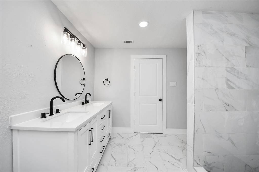 This bathroom features a modern design with a double vanity, sleek black fixtures, and a round mirror. The space is accented with elegant marble tiles and a neutral color palette, creating a clean and sophisticated atmosphere.