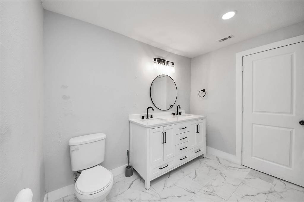 This bathroom features a modern design with a sleek white vanity, dual sinks, and black fixtures. It has a round mirror, ample storage, and is accented with elegant marble-patterned floor tiles.