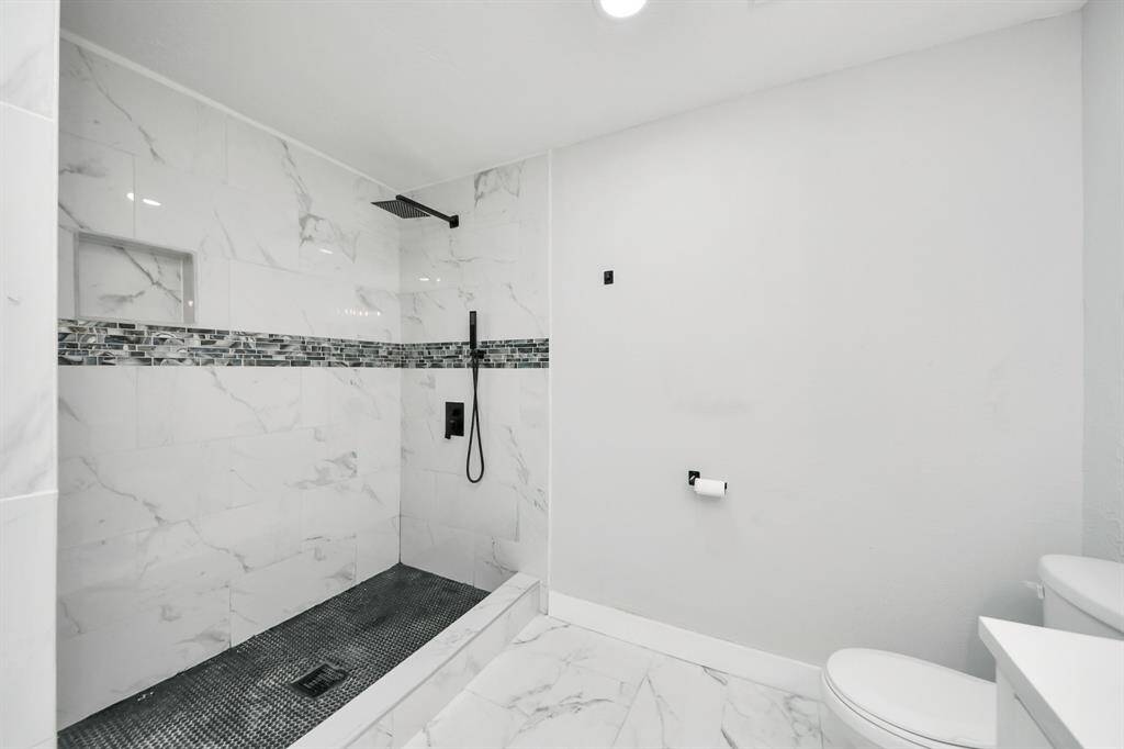 This bathroom features a spacious, modern design with a walk-in shower adorned with marble-like tiles and a sleek black showerhead. The minimalist look is complemented by a subtle mosaic tile accent, providing a contemporary and elegant touch.