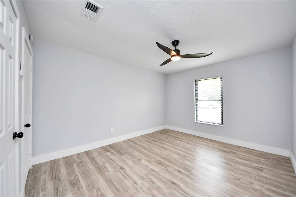 This photo shows a bright, unfurnished room with light wood flooring, a modern ceiling fan, and a single window with blinds. The walls are painted a soft gray, enhancing the room's airy feel.