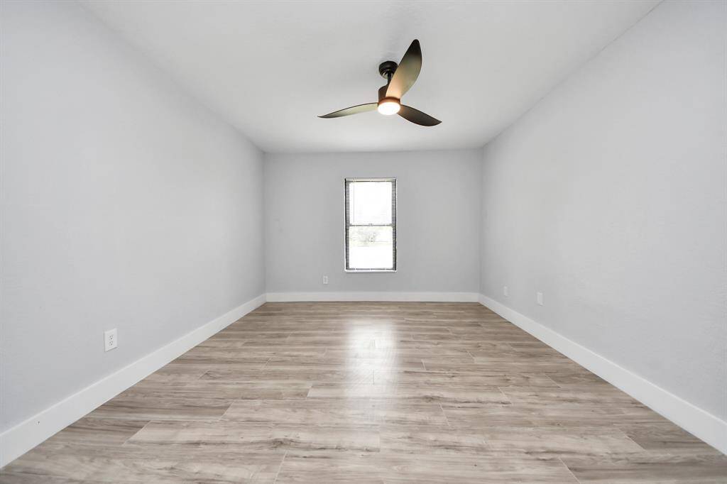 This room features a spacious layout with light gray walls and wood-style flooring. It has a ceiling fan and a window allowing natural light to fill the space, creating a bright and airy atmosphere.