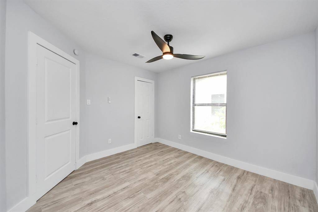 Bright, minimalist bedroom with light wood flooring, neutral walls, a modern ceiling fan, and a single window for natural light. Two doors provide access to closets or adjoining spaces.