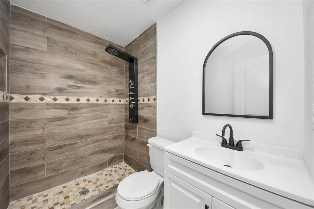 This bathroom features a modern walk-in shower with wood-look tile and decorative accents. It includes a sleek black shower panel, a white vanity with a black faucet, and an arched mirror, all set against a clean, bright white wall.