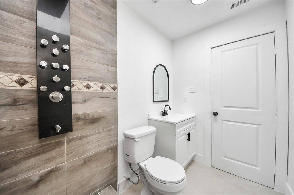 This modern bathroom features a sleek shower panel with multiple jets, elegant wood-look tile, a compact vanity with a black faucet, and a clean white color scheme.