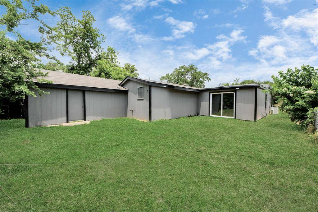 This photo showcases a single-story home with a modern exterior featuring gray siding and a flat roof. The house is set on a spacious, well-maintained lawn with mature trees providing a serene atmosphere. Large sliding glass doors suggest ample natural light and indoor-outdoor living potential.
