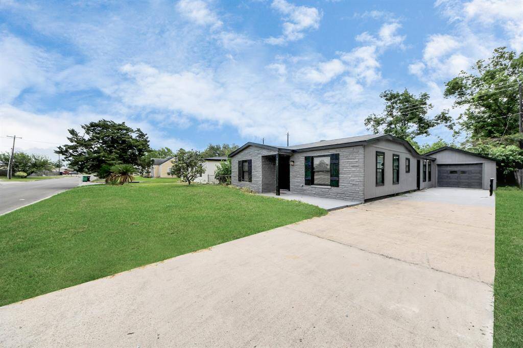 This photo shows a modern, single-story home with a light gray exterior and a large driveway leading to a detached garage. The property features a spacious front lawn and is situated on a corner lot with clear skies above.
