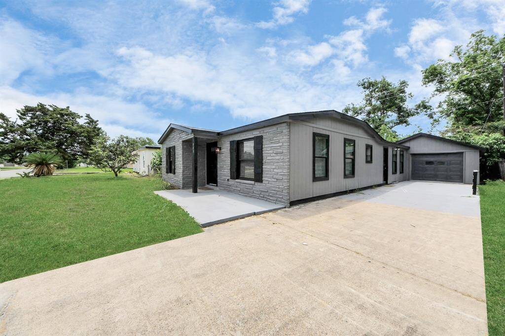 This photo shows a modern, single-story home with a gray exterior and stone accents. It features a spacious driveway leading to an attached garage, surrounded by a well-maintained lawn and mature trees, offering a welcoming and comfortable atmosphere.