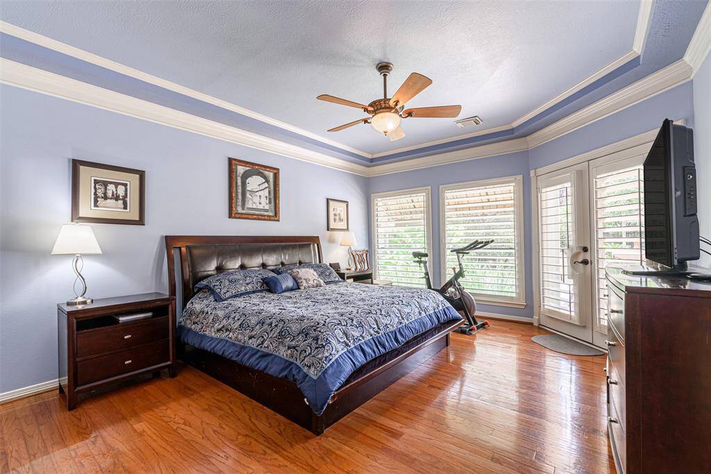 This primary suite features a complemented tray ceiling and ceiling fan. It has two windows providing ample natural light, and wood floors. Double doors lead to the pool area.