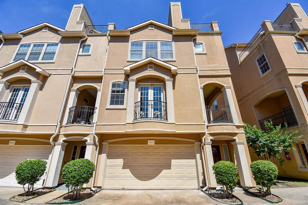A modern townhouse with a light-color exterior, featuring multiple stories and attached garages. The architecture includes arched balconies and decorative trim. The property is situated in a clean, paved area with neatly trimmed bushes, under a clear blue sky.