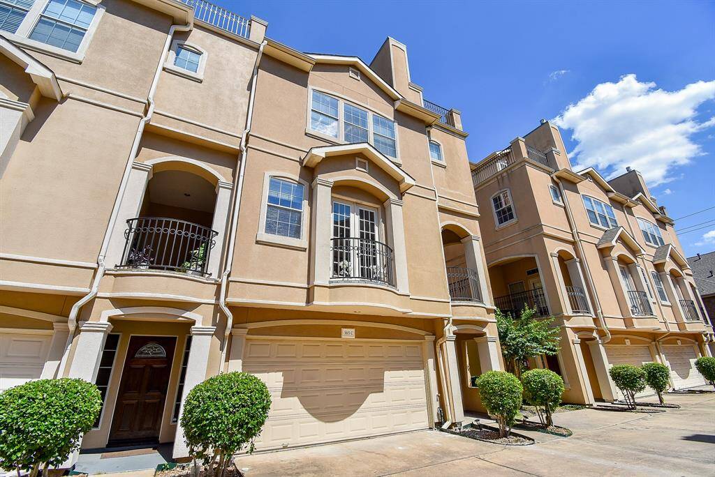 A row of modern townhouses with a stucco exterior and decorative balconies. Each unit features a two-car garage and a neatly maintained driveway. The architecture is contemporary, with clean lines and large windows, offering appealing facade.