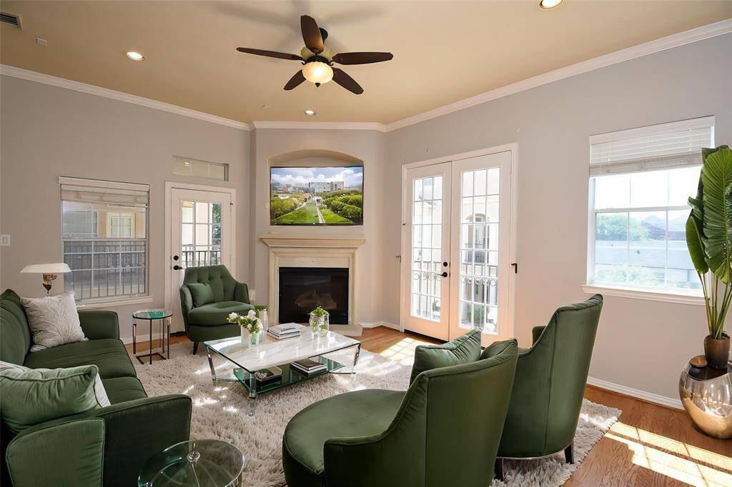 Bright living room with hardwood floors, a cozy fireplace, and French doors leading to a balcony. Features crown molding, recessed lighting, and a ceiling fan. Ideal for comfortable living.
