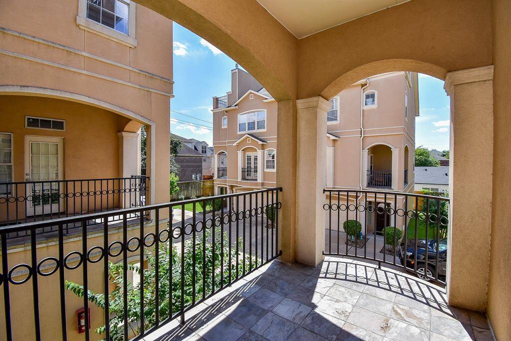 This photo showcases a cozy, covered balcony with tiled flooring and a wrought iron railing. The space features a glass-paneled door and a window, and a view of neighboring buildings. Ideal for a small outdoor seating area.