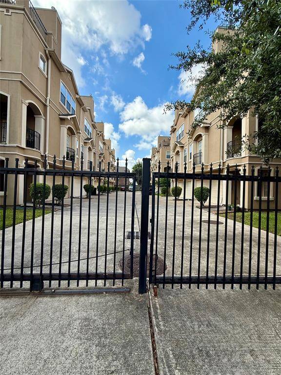 This is a three-story townhouse with a beige exterior and white trim, featuring two balconies and a two-car garage. The home has a clean, modern look with well-maintained landscaping, including small shrubs at the entrance.
