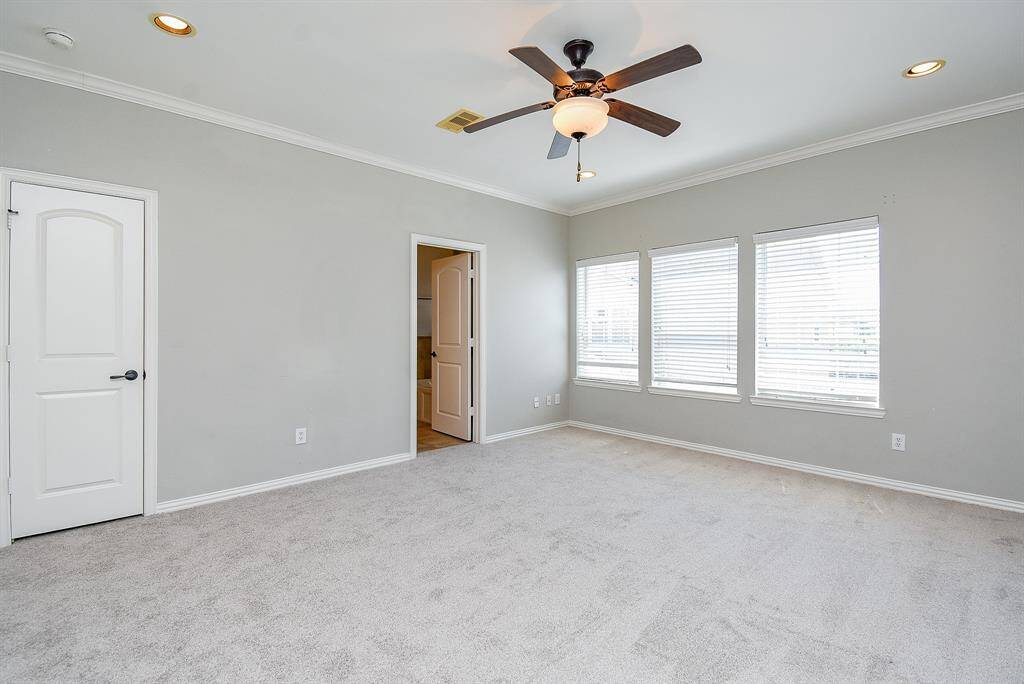 This spacious room features soft carpet, neutral gray walls, and ample natural light from three large windows. It includes recessed lighting and a ceiling fan for comfort and style.
