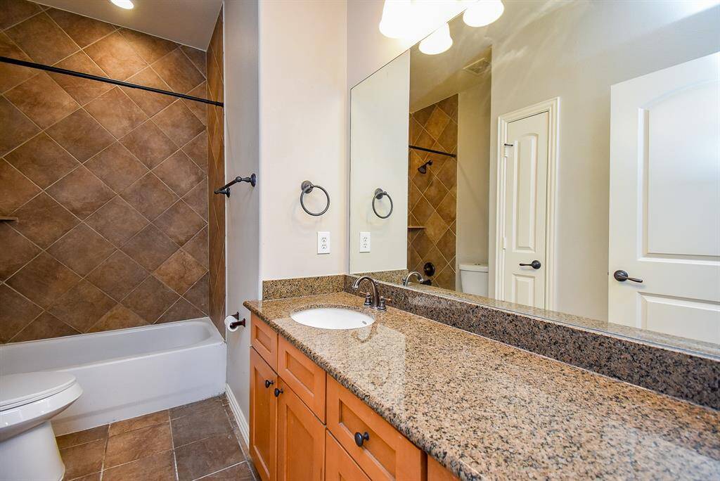 This photo shows a modern secondary bathroom with a granite countertop, wooden cabinetry, and a single sink. It opens to the hallway. The area is well-lit and has a clean, neutral color palette.