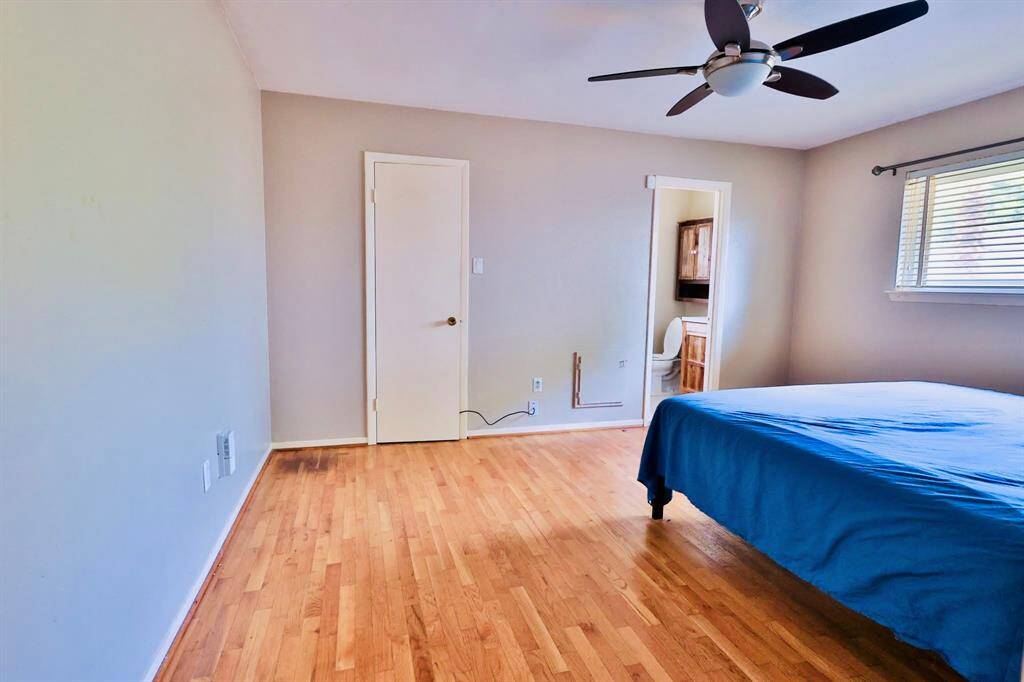 Bright bedroom with hardwood floors, ceiling fan, and a window for natural light. Features an attached bathroom and neutral wall colors, offering a cozy and functional space.