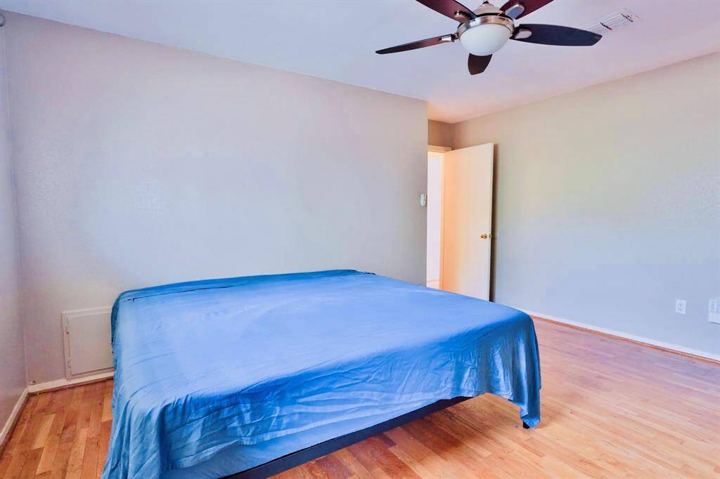 This photo shows a spacious bedroom featuring hardwood floors, neutral walls, and a ceiling fan. The room is well-lit, with a simple bed setup and a partially open door, suggesting access to another area or hallway.