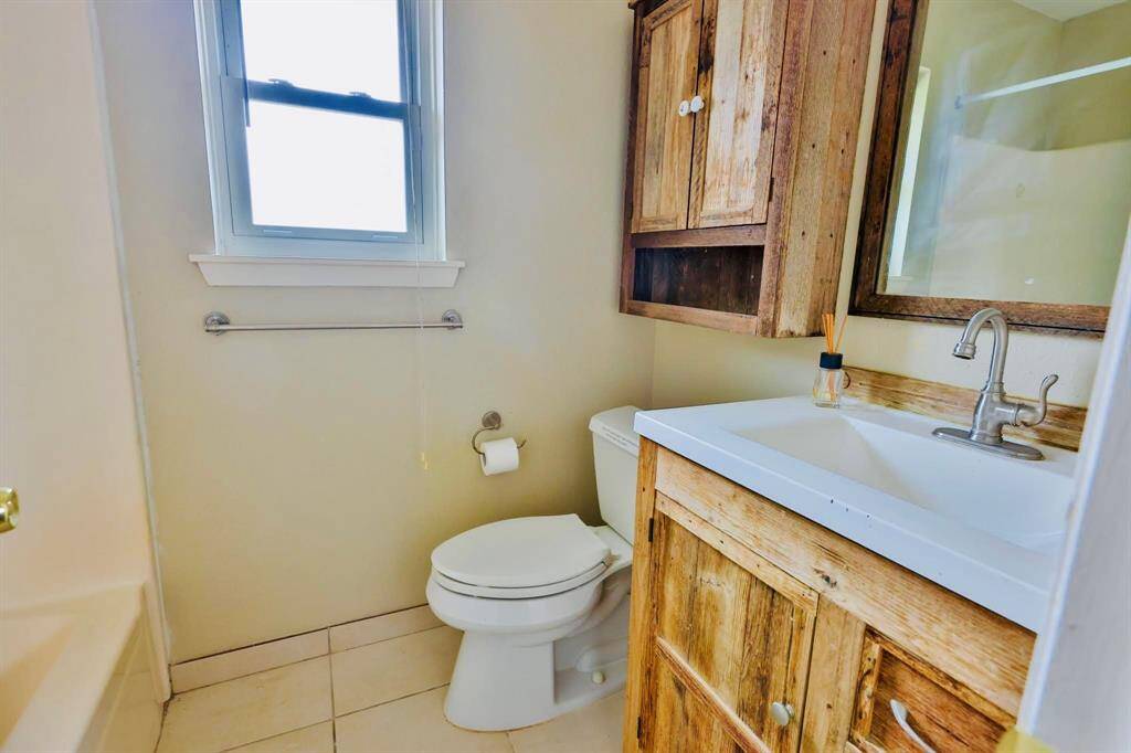 This bathroom features a rustic charm with wooden cabinetry and a matching mirror frame. It includes a toilet, sink, and a bathtub. The room is well-lit with natural light from a window above the toilet, enhancing the bright, welcoming atmosphere.