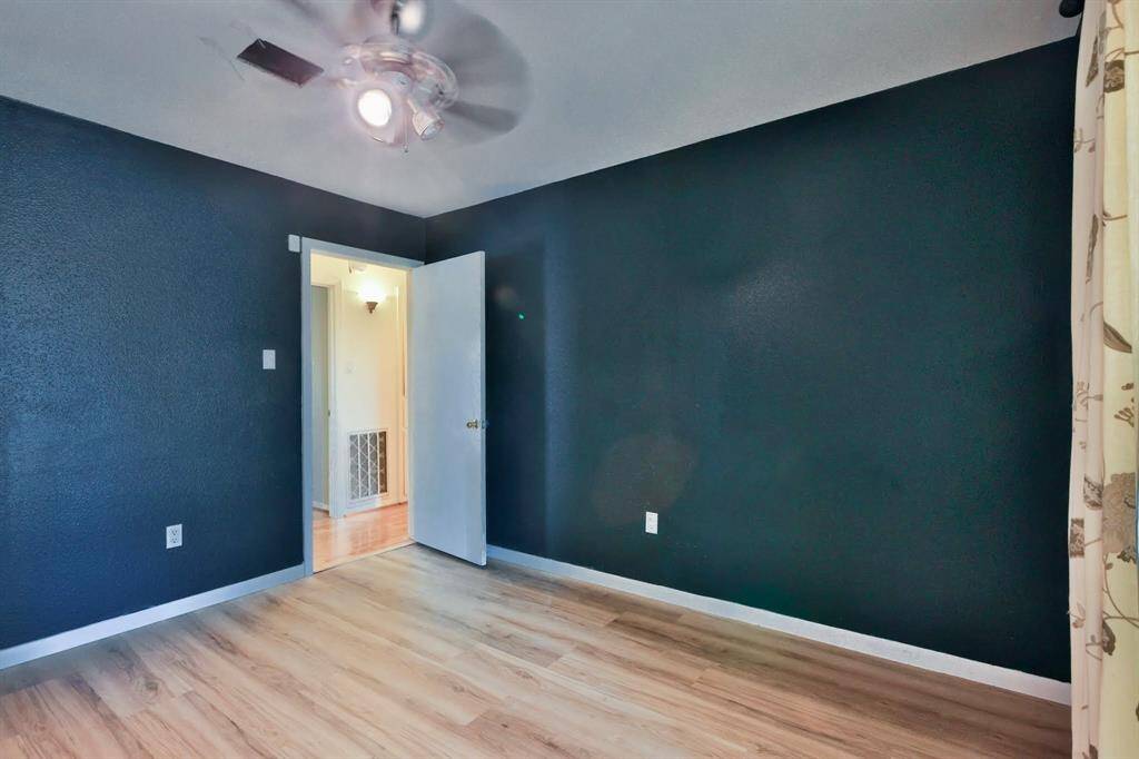 This room features dark blue walls, light wood flooring, and a ceiling fan. It has an open doorway leading to a brighter area, and a window with patterned curtains, providing a cozy and modern feel.