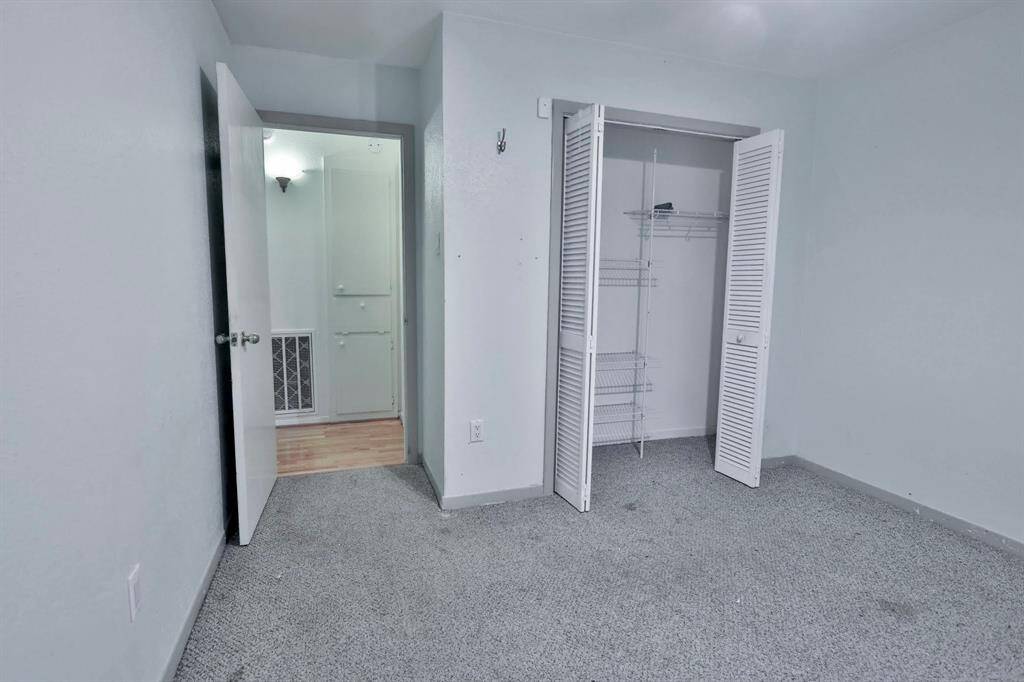 This photo shows a small room with light-colored walls and carpet flooring. There's an open closet with white louvered doors and wire shelving. A door leads to a hallway with wooden flooring and simple wall lighting.