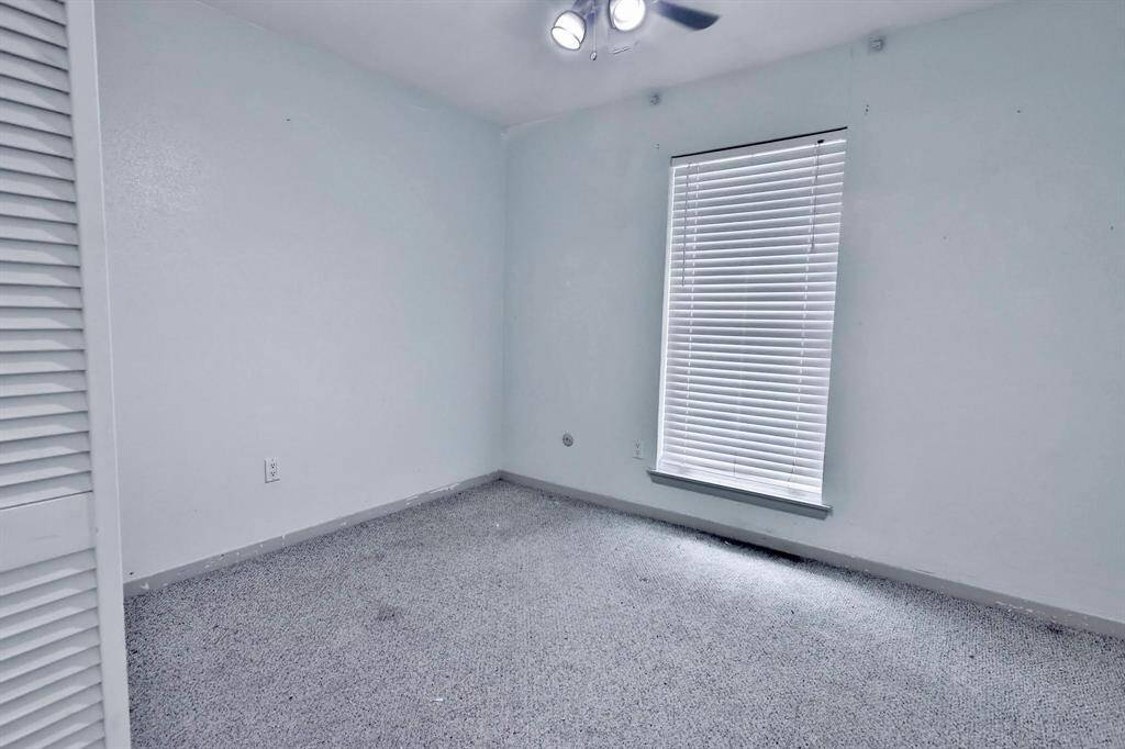 This is a small, empty room with light-colored walls and carpeted flooring. It features a window with blinds and a ceiling fan, offering natural light and ventilation.