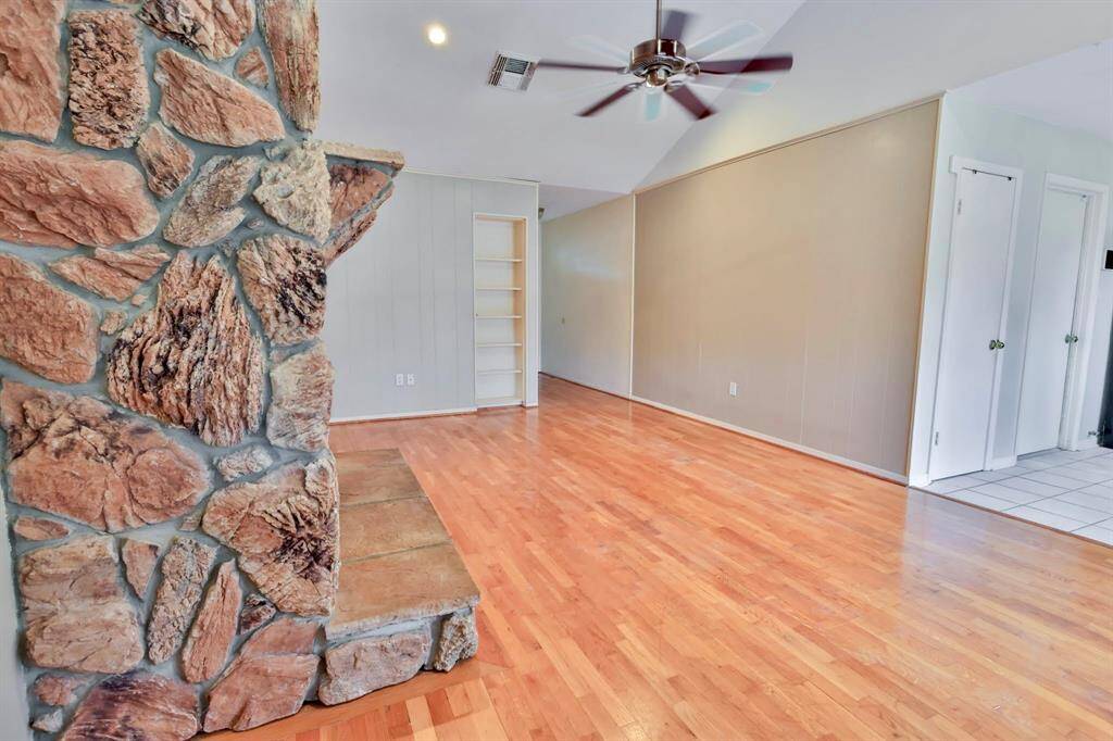 This room features a striking stone fireplace, beautiful hardwood floors, and a ceiling fan. It offers an open layout with built-in shelving and leads to a tiled entryway.