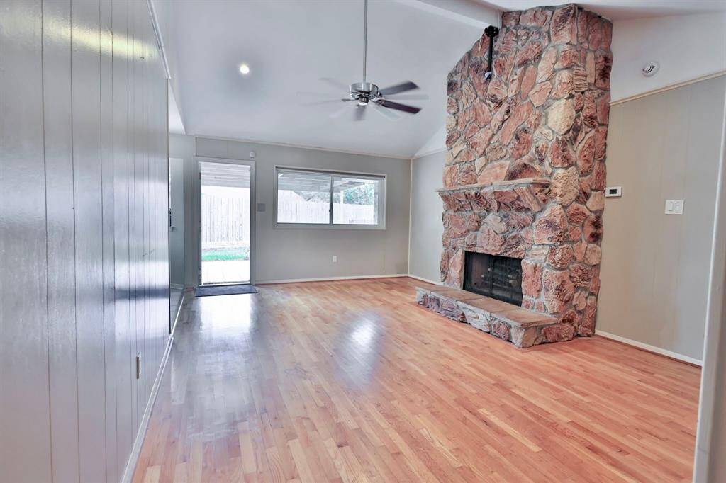 This room features a cozy stone fireplace, hardwood floors, and a ceiling fan. It has natural light from large windows and a door leading to the backyard, creating an inviting and warm atmosphere.